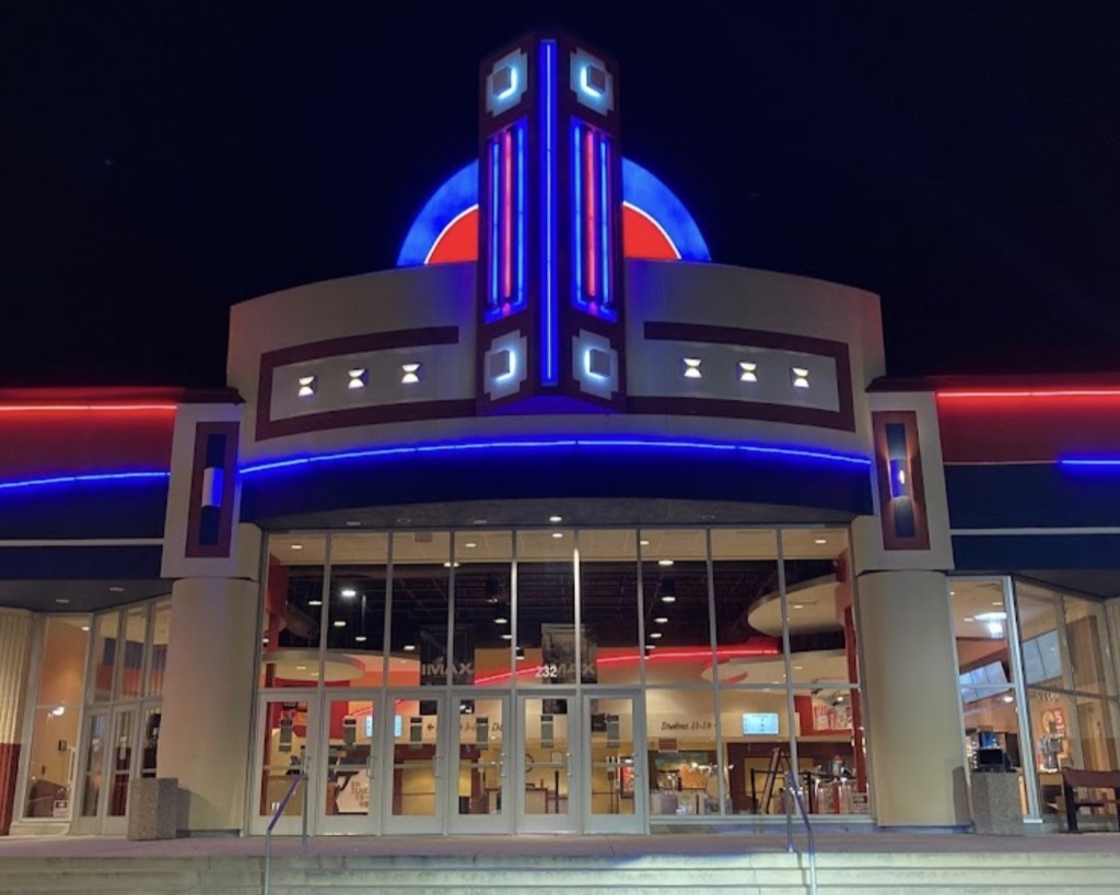 A nighttime shot of the Savoy 16 + IMAX movie theater. There is a black sky and the theater is lit up with red and blue neon lights. There are lights on inside and people in the lobby visible through the wall of doors and windows.