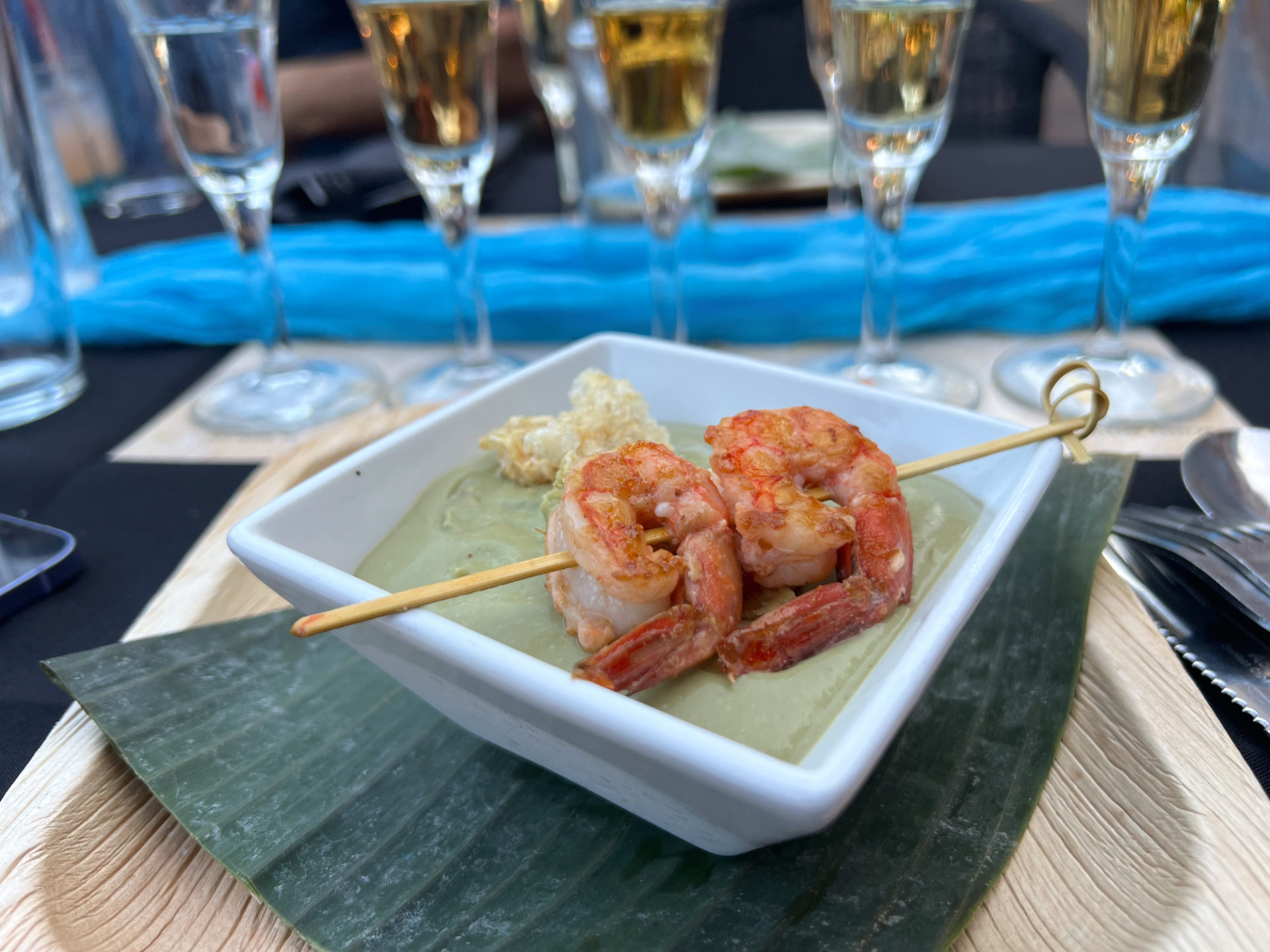 A white square bowl has two shrimp skewered on a bamboo pick over a chilled avocado soup.