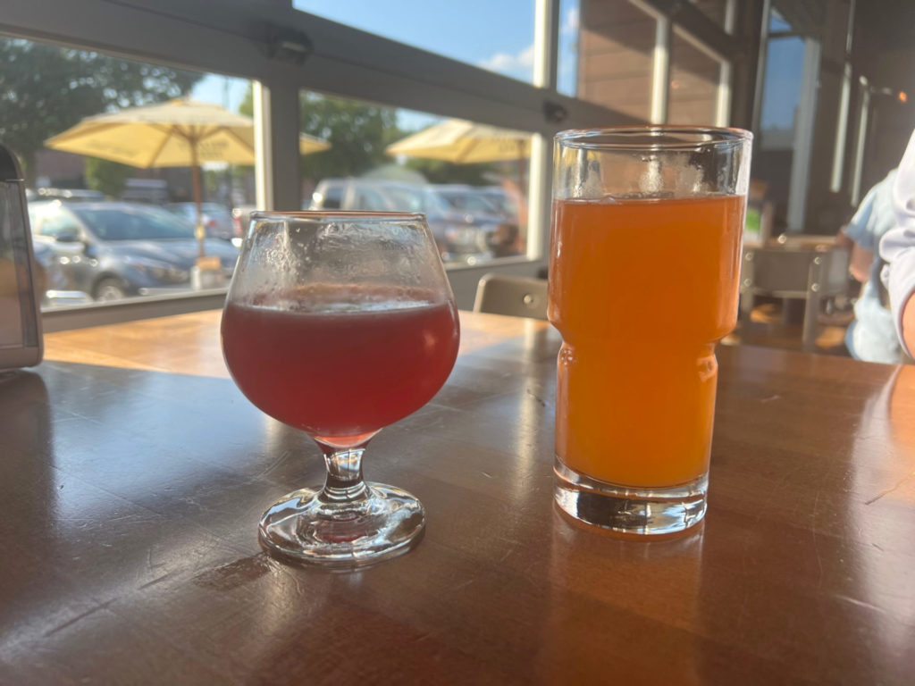 On a brown table, there are two drinks: a red cocktail called Paper Plane and an orange beer. Photo by Alyssa Buckley.