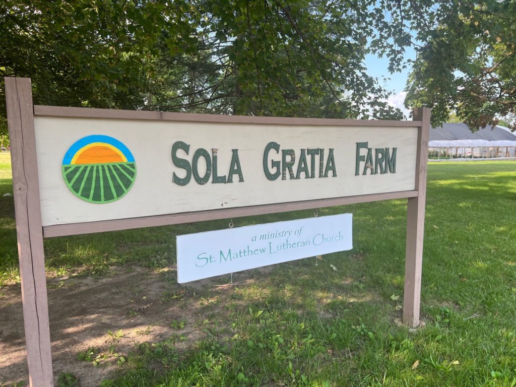 A long, rectangular wood sign. It's painted light beige, and has the words Sola Gratia Farm in green block letters. 