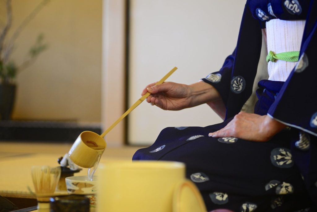 A person in traditional Japanese clothing is kneeling and stirring tea with a long wooden stick.