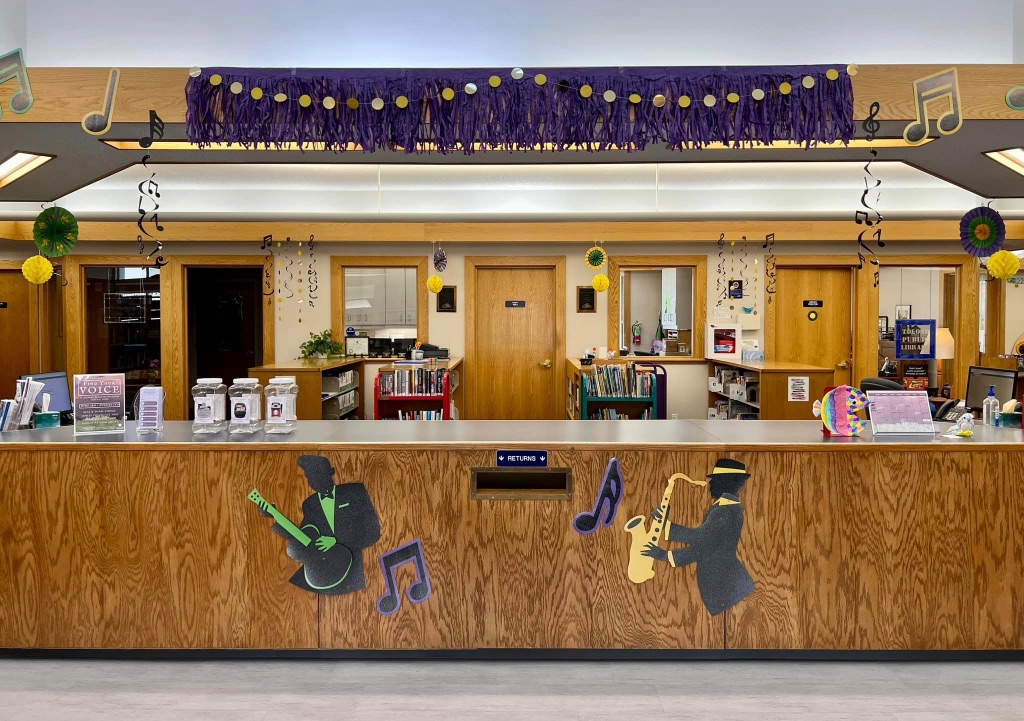 A picture of a welcome desk at the Tolono public library. It is all wood with a white counter. There are cut outs of music notes and jazz musicians decorating the facade. 