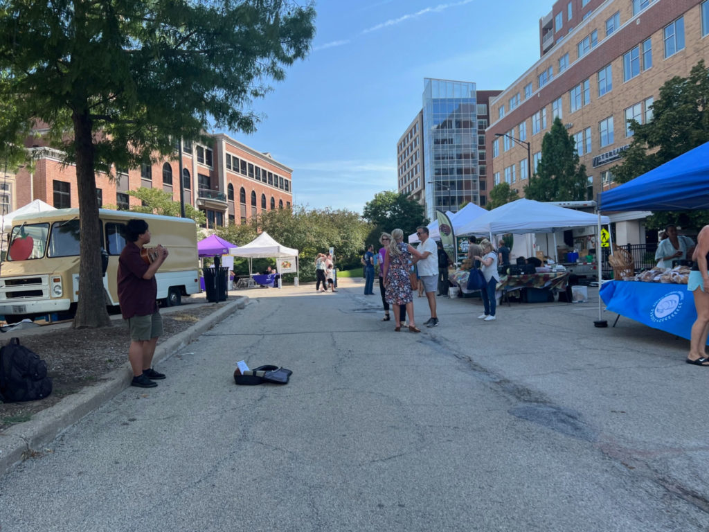 At the Tuesday market in Champaign, there is a musician playing in the parking lot while people shop The Land Connection's Champaign farmers market. Photo by Alyssa Buckley.