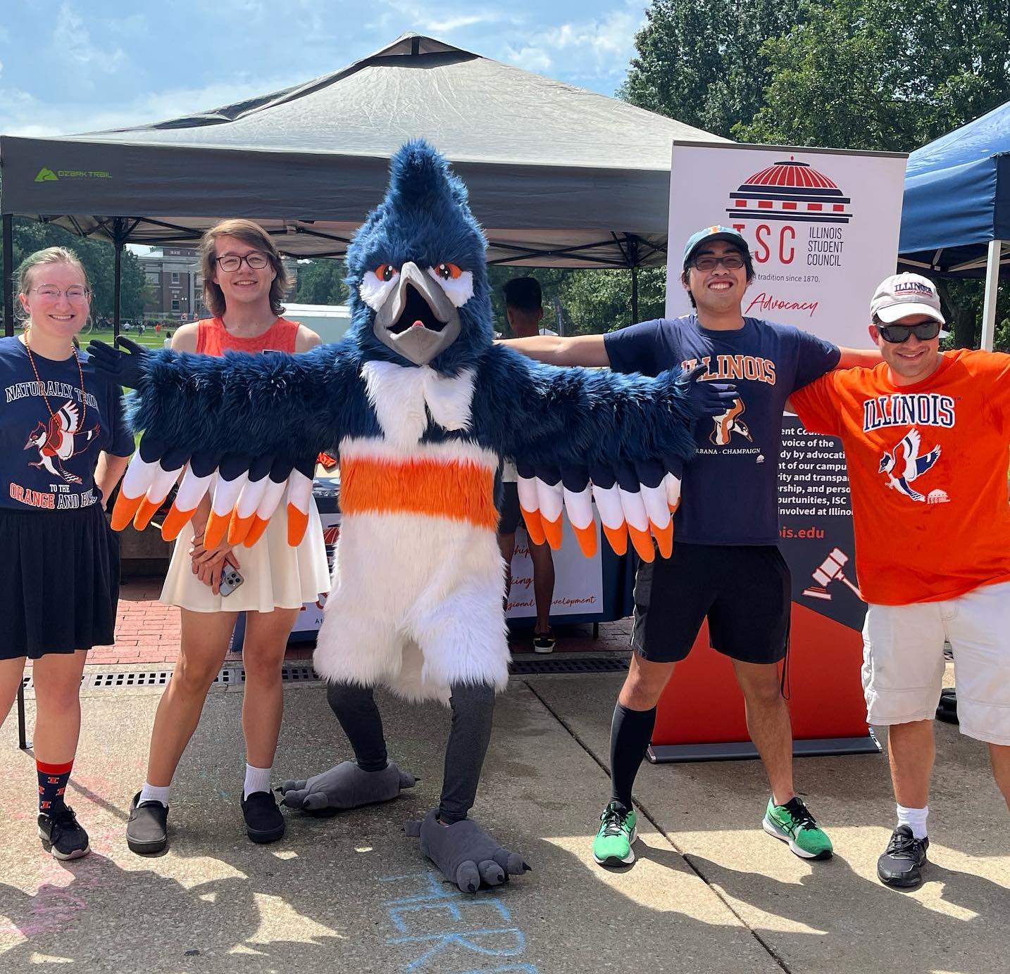 A kingfisher mascot stands with arms spread open. On each side are two people, wearing blue and orange t-shirts. The kingfisher costume is a blue, white, and orange bird. They are standing outside in front of a tent.