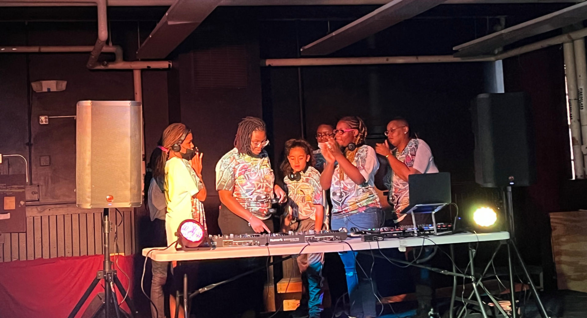 Six Black young girls and women stand around a table covered in DJing equipment. They all appear to be wearing the same t-shirt. Some of the people are clapping.