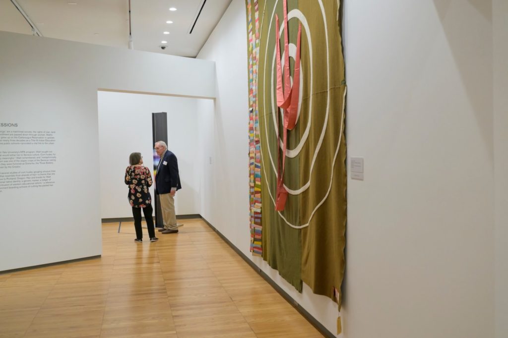 patrons stand in Krannert Art Museum looking at the Marie Watt exhibit. One of her pieces is visible on the side. It is a large fabric piece in green-yellow with concentric circles. 