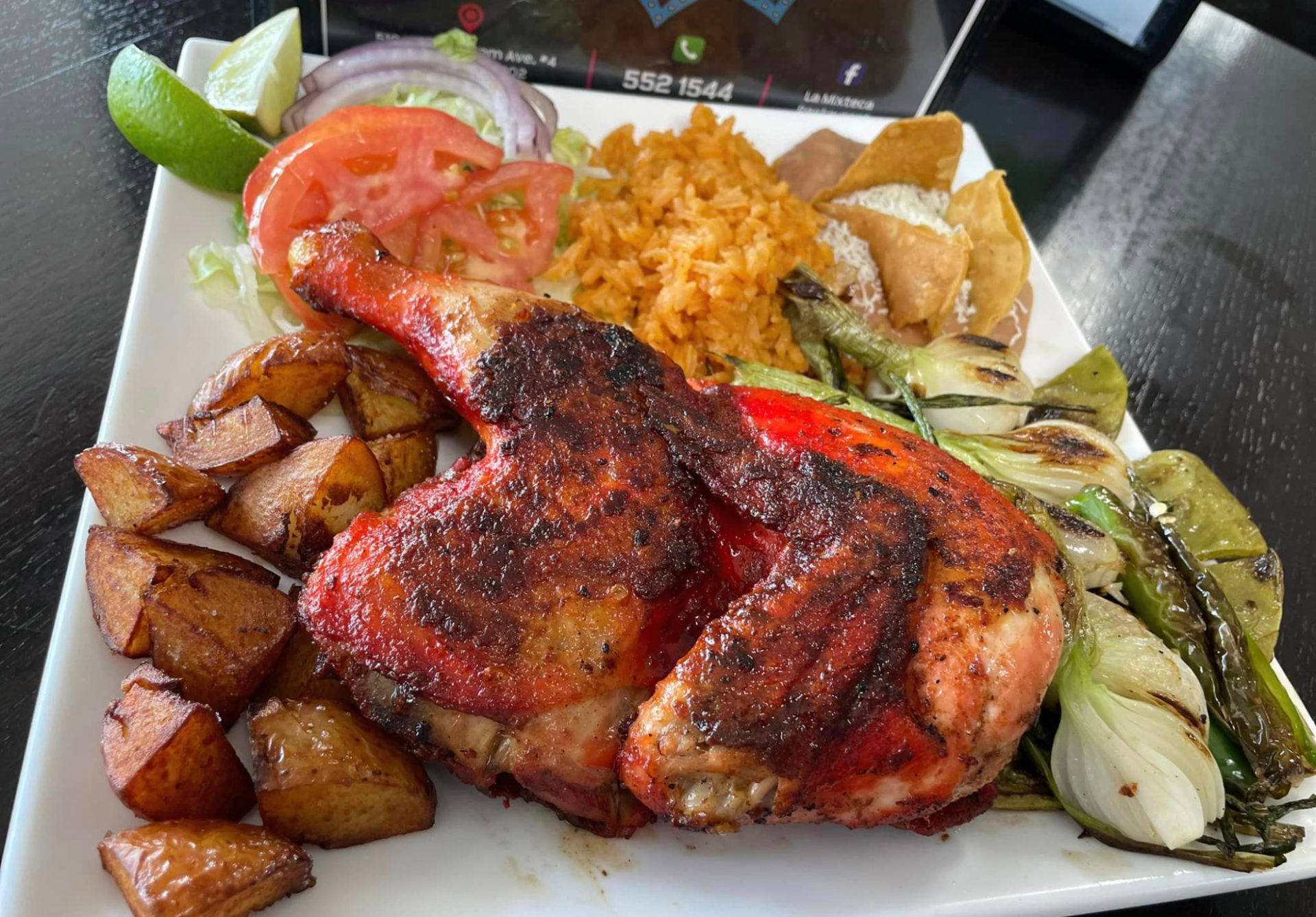 A dish of grilled chicken thighs with wings attached, roasted potatoes, grilled onions, rice, and beans served on a white plate at La Mixteca.