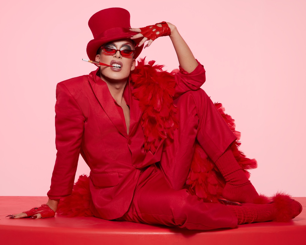 Alaska 5000 sits on a red cushion against a pink background, she wears a red jacket, pants, tophat, and sunglasses. She has red fingerless gloves and one hand on her hat. She is white with very long blonde hair.