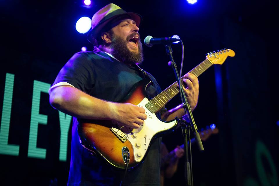 a photo of a musician playing an electric guitar on stage. The musician is wearing a black t-shirt and a brown hat. The guitar is a Fender Stratocaster with a sunburst finish. The background is a stage with blue and purple lighting. The musician is singing into a microphone.
