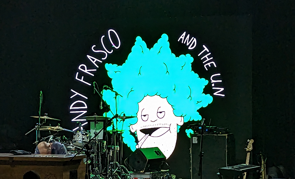 The stage is set for a performance, illuminated by white and blue lights that cast an ethereal glow. A large screen dominates the backdrop, featuring a cartoon image of a man with striking blue hair and a vibrant green shirt. The text above the image announces “Andy Frasco and the UN”, hinting at the act to follow. Musical instruments, including a drum set, are poised on the stage, ready to bring music to life. The background is swallowed in darkness, allowing the stage and its setup to take center stage in anticipation of the performance.