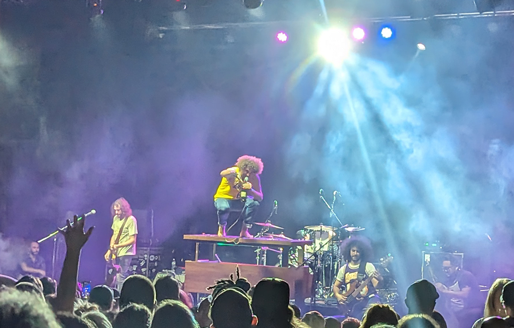 A rock concert is in full swing, the stage awash with the glow of blue and white lights. The lead singer, clad in a yellow shirt, commands the stage from atop a piano, microphone in hand. To their side, a guitarist strums a vibrant red guitar, while the bassist plucks at the strings of a blue bass guitar. Behind them, the drummer keeps time on a drum kit complete with cymbals. The audience, visible in the foreground, is caught up in the energy of the performance, their hands raised high in the air. The backdrop to this lively scene is a large screen and various pieces of stage equipment.