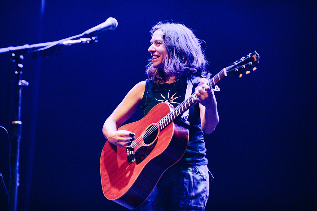 A white woman on a stage playing an acoustic guitar. The individual is dressed in a black tank top adorned with a white design. The guitar they’re strumming is a reddish-brown color, secured with a black strap. The backdrop is a deep blue hue, with stage equipment and a microphone stand discernible in the background. A spotlight illuminates the scene.