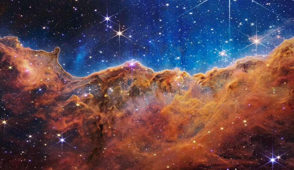 An image of a star nursery. Giant orange clouds of gas take up the lower half of the image with dark blue skies and star clusters.