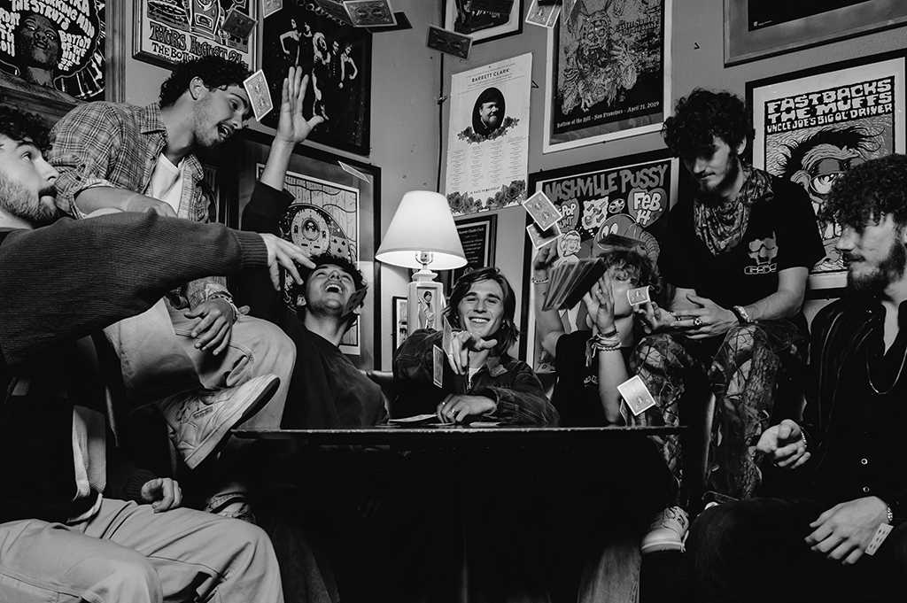 This is a black and white photo of a group of people in a room with posters and other decorations on the walls. The people are sitting and standing around a table with a lamp on it. The posters on the walls are of bands and musicians, including “Nashville Pussy” and “The Fastbacks”. There is a guitar hanging on the wall in the background. The room appears to be a music or band practice space.