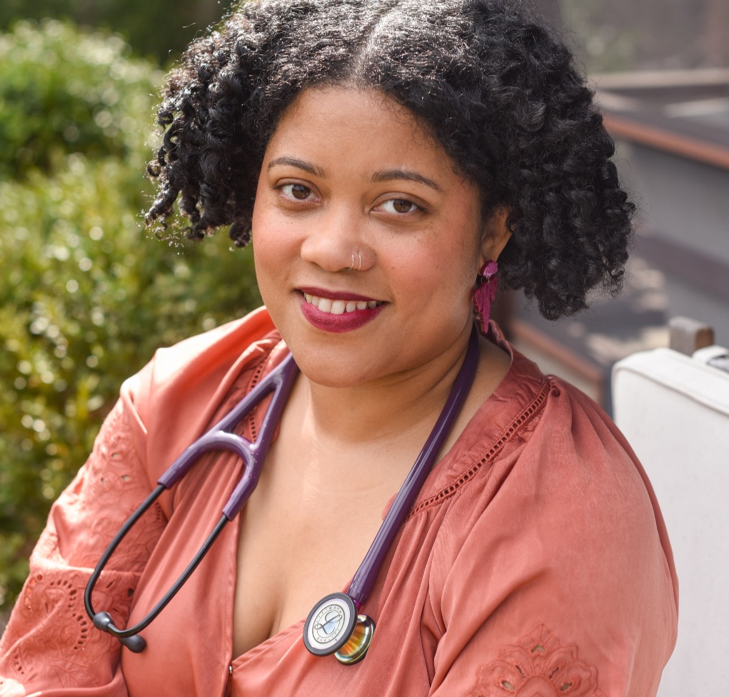 A brown woman with short curly hair wears a pink long sleeve blouse and a stethoscope she is smiling at the camera.