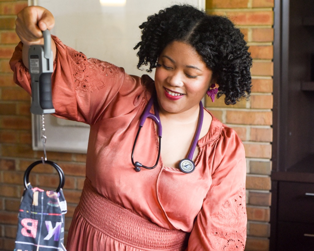 A brown woman with short curly hair wears a pink long sleeve blouse and a stethoscope she is holding a cloth suspended from a hook, used to weigh babies.
