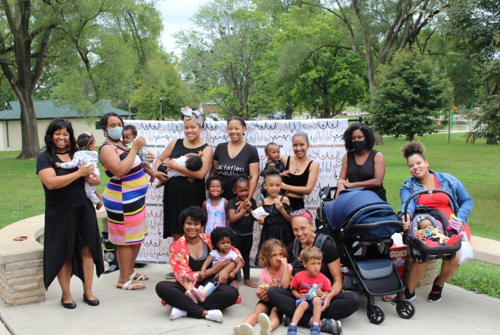 A group of black woman and babies of various ages stand together in a park with green leafy trees behind them. 
