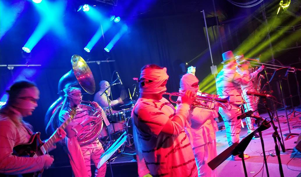 A band is performing on a stage illuminated by blue and purple lights. The band members are dressed in red and white striped outfits and hats. They are playing various instruments, including a guitar, a sousaphone, a drum set, and a trombone. The background consists of a stage with curtains and lighting equipment.