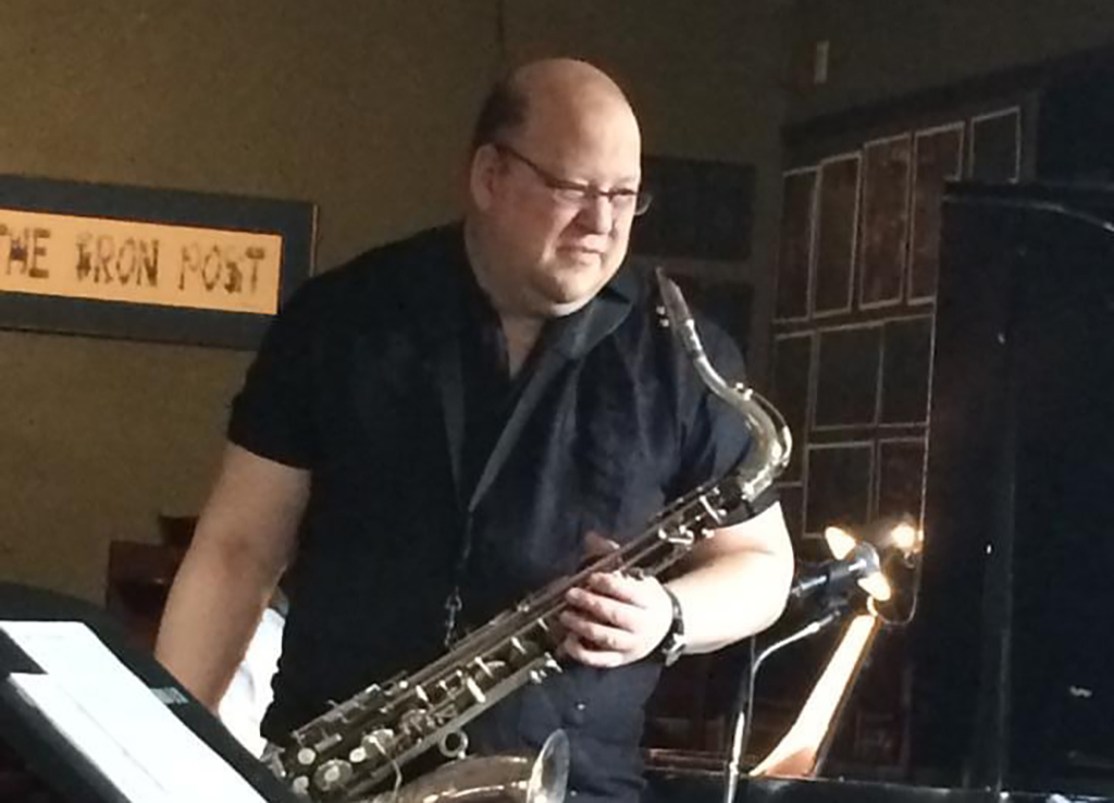 A person is playing a saxophone in a dimly lit bar. They are wearing a black shirt and standing in front of a music stand. The saxophone is brass-colored and the person is holding it with both hands. The background consists of a brick wall and a sign that reads “The Iron Post”.