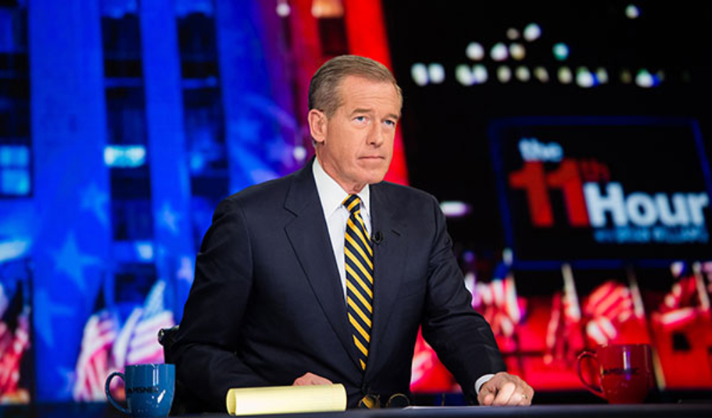 Journalist Brian Williams, a middle aged white man, wears a dark suit with a white shirt and yellow and navy striped tie. He sits behind a desk on the set of the TV show The 11th Hour with Brian Williams.