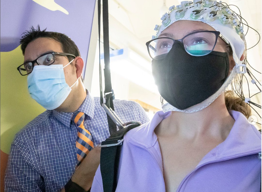 A man in a surgical mask is standing alongside a woman, also wearing a mask, who has a cap with wires coming out of it on her head.