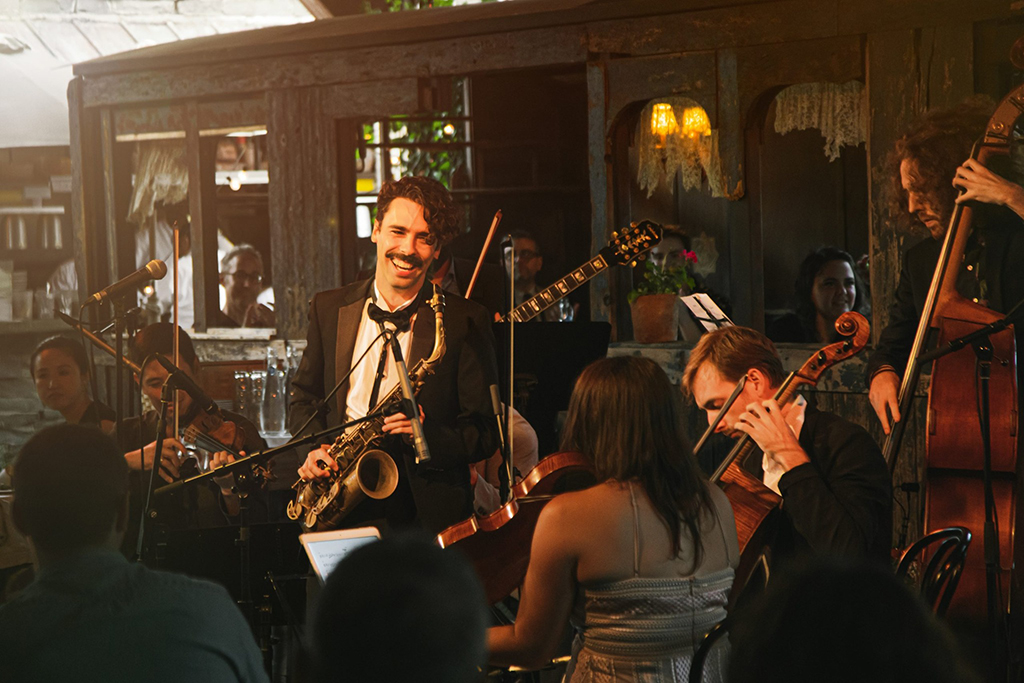 A lively jazz band is performing in a dimly lit restaurant. The band consists of a saxophonist, a guitarist, a bassist, and a cellist. The guitarist and bassist are playing their instruments, while the cellist is tuning theirs. The restaurant has wooden walls and a chandelier hanging from the ceiling. In the background, people are sitting at tables, enjoying the music. The atmosphere is intimate and full of life.