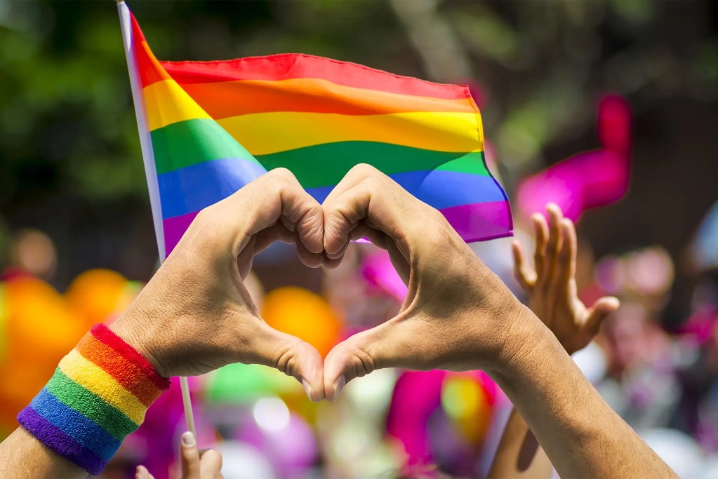 Two hands make a heart, on the wrist is a rainbow sweatband and there is a blurry crowd and a rainbow striped flag in the background.