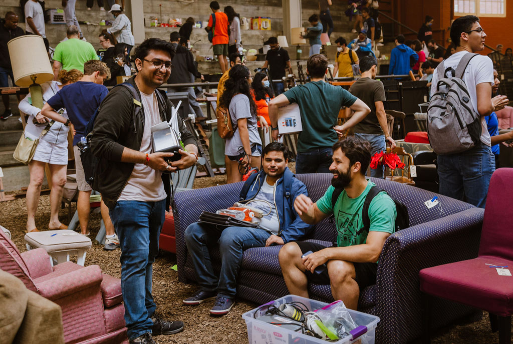 A huge group of people are in a warehouse full of furniture. There are two brown men sitting on a blue couch and one brown man, holding a light standing in front of them. They are all smiling. 