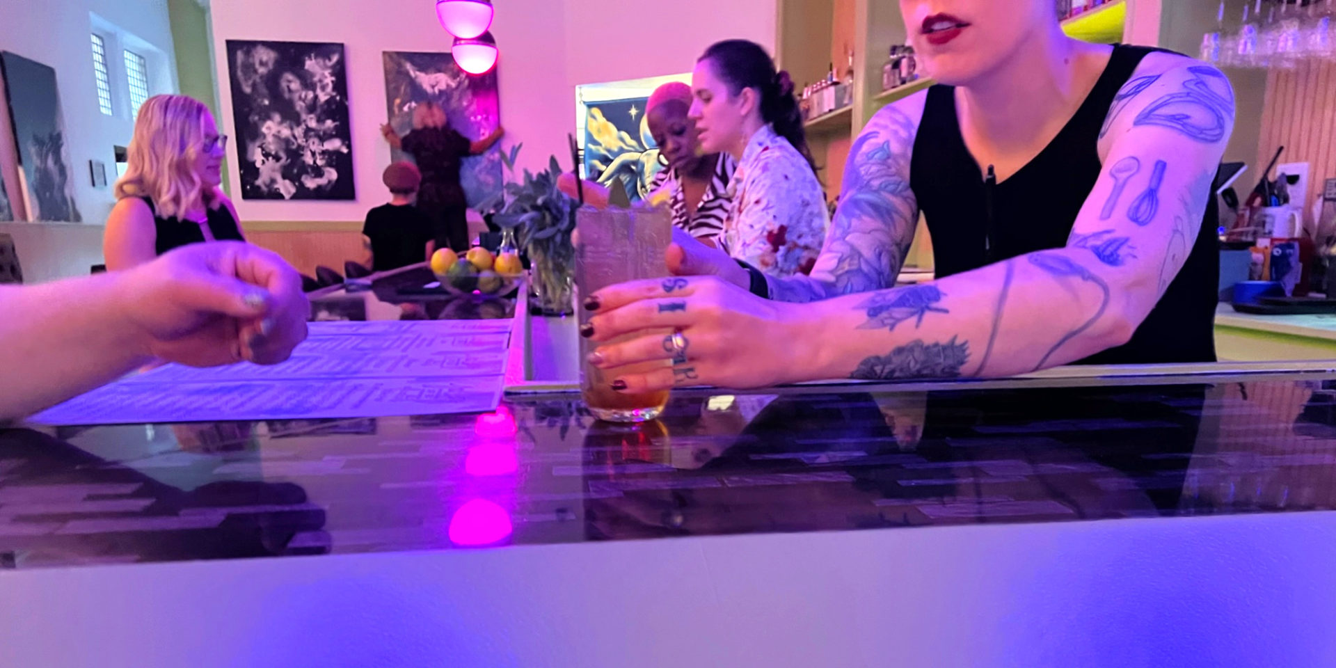 A bartender at Gallery Art Bar serves a mocktail to a white patron on the left. Two other bartenders discuss drinks behind the bar, and in the background an artist hangs her painting on the wall.