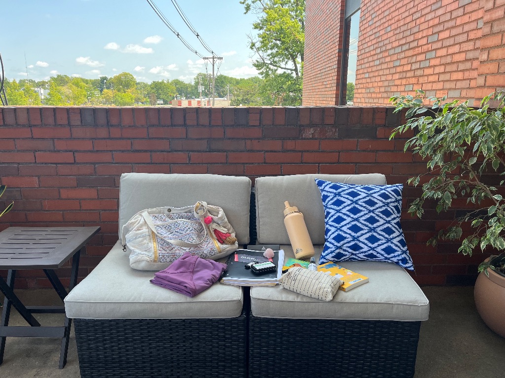 A close up shot of a white couch. On it are items from Lola's bag. Some of them include a purple folded yoga tank top, a white bag with a faded design embroidered, a peach water bottle and a book with a yellow cover.
