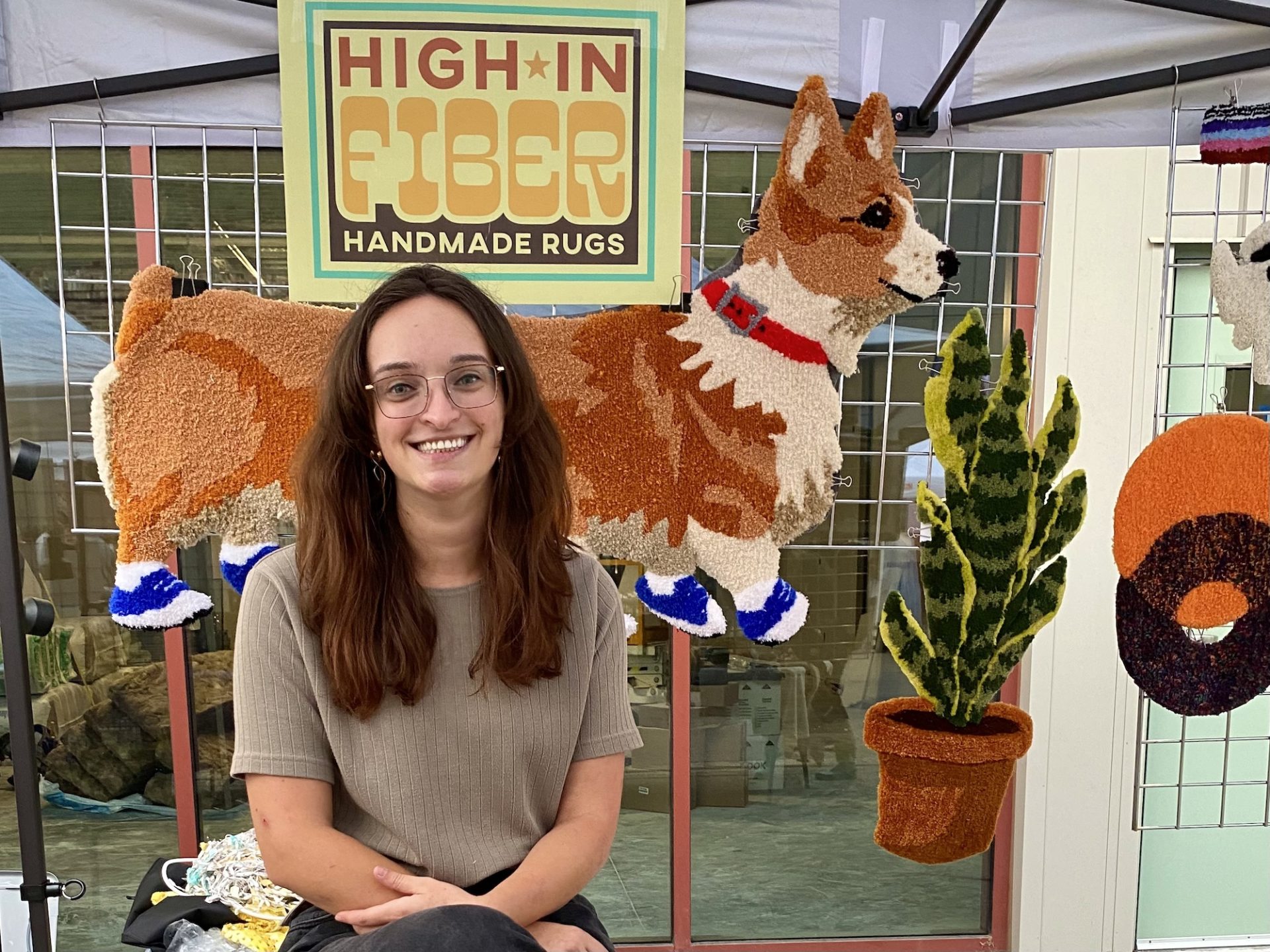 Brittany Heyen, a young white woman with glasses and long brown hair, sits in front of her booth. Her High in Fiber Rug sign is above along with the rug corgipillar