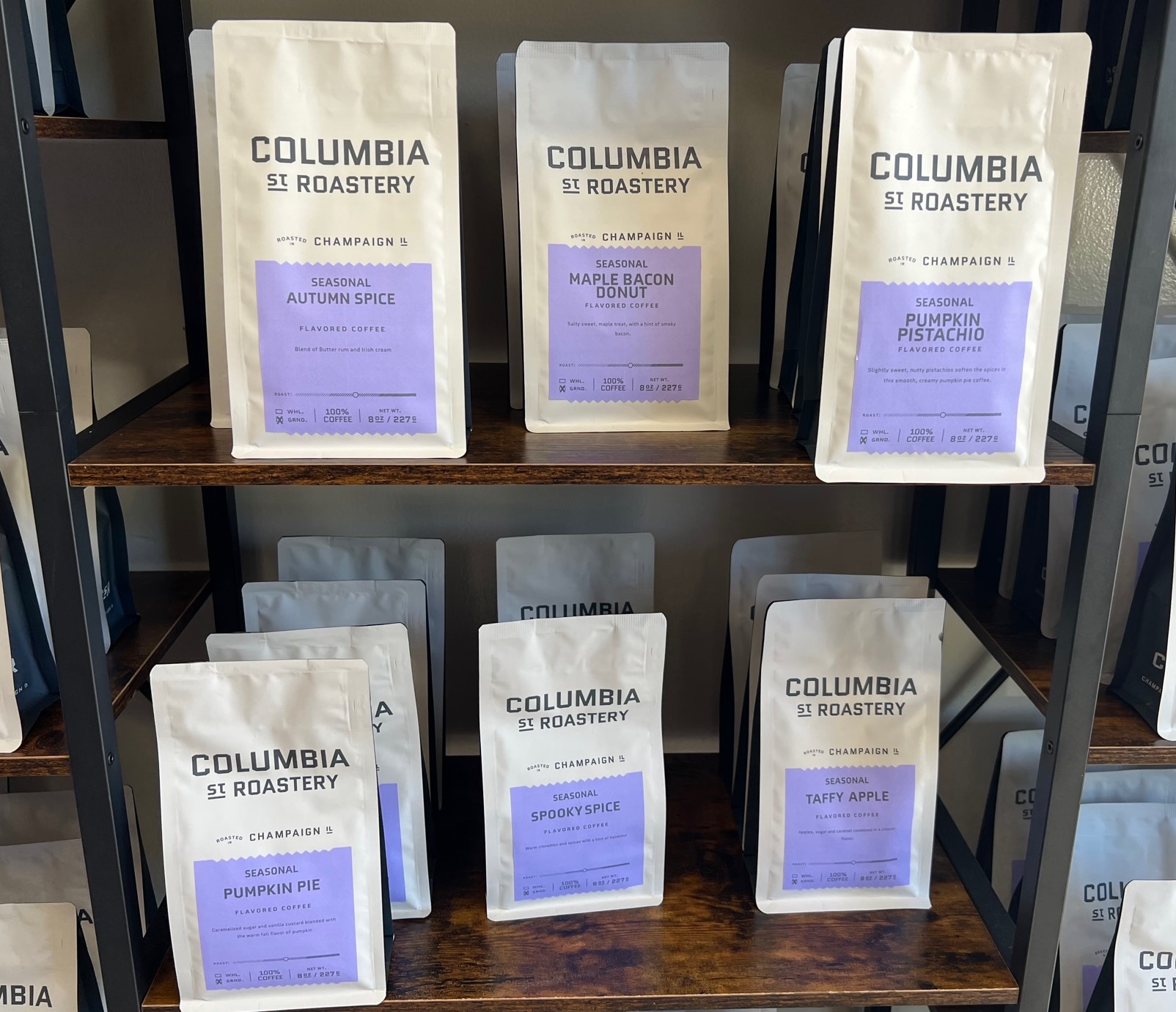 Bags of coffee are lined up on wooden shelves. The packages are off-white with a purple label.