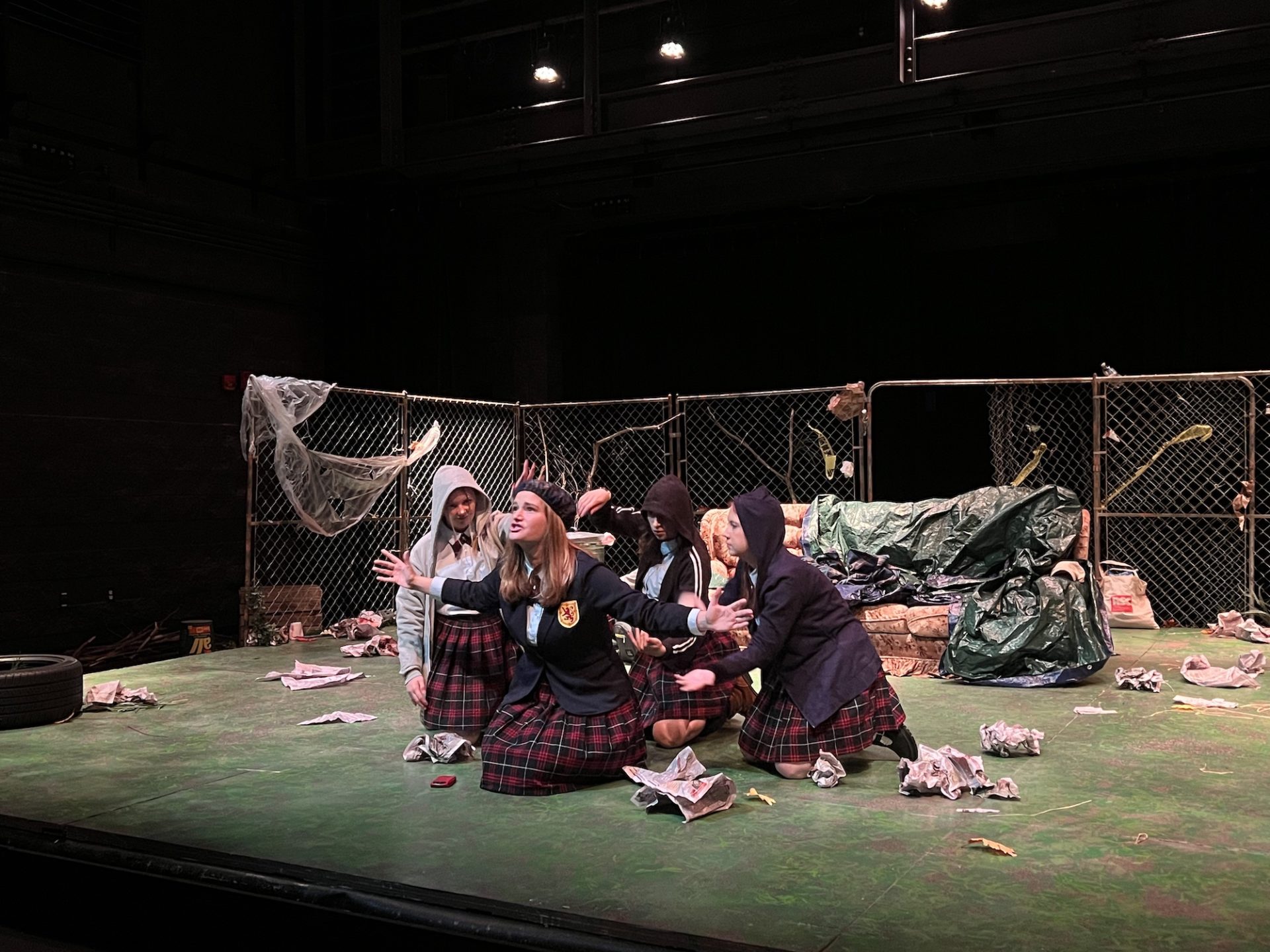 Actresses wearing school uniforms sit on the floor of the stage. Lady Macbeth is in the center with the three witches surrounding her as she gives a passionate monologue. The set looks like an abandoned lot, with a dirty old couch and garbage everywhere.