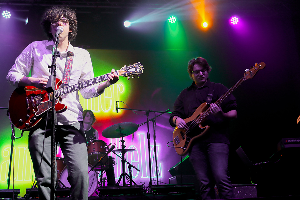 A band is in the throes of a performance on a stage bathed in colorful lighting. The backdrop is a vibrant mix of purple and green, with the band’s name emblazoned across it. The ensemble consists of two guitarists and a drummer. The guitarist on the left strums a red electric guitar, dressed in a white shirt and a black tie. In the background, the drummer is engaged with a white drum kit. On the right, another guitarist is visible. The stage is awash with lights in hues of purple, green, and pink.