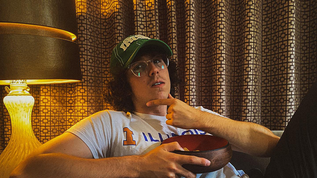 A person is sitting on a couch with a bowl in their lap. They are wearing a white t-shirt with “I - Illinois” written on it and a green baseball cap with "Irish" on it. The bowl is brown and red and appears to be nearly empty. The background consists of a brown curtain with a geometric pattern and a lamp with a beige lampshade.