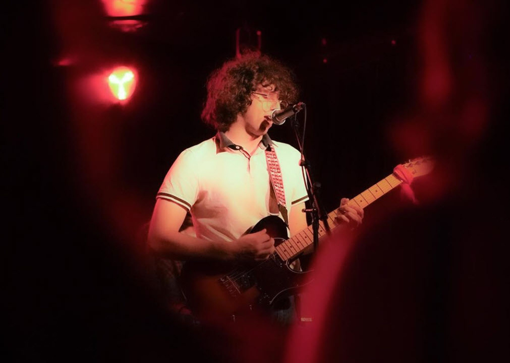 A musician is on stage under red lighting, playing a black electric guitar. They are dressed in a white shirt with black trim. The background is dark, and a silhouette of a microphone stand is visible. The perspective is from someone in the audience, as the back of a head can be seen in the foreground.