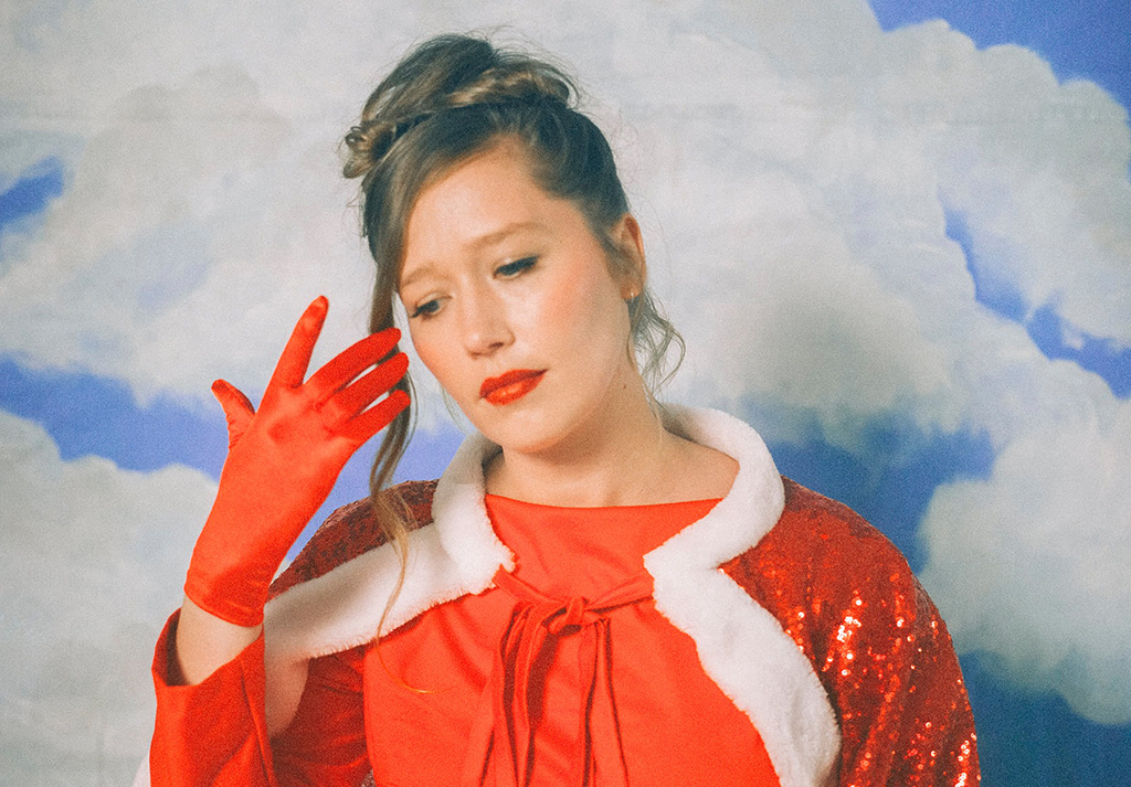 a person striking a pose against a backdrop of a blue sky scattered with white clouds. They are dressed in a vibrant red dress that features a white fur collar, adding an element of elegance. Complementing the dress are matching red gloves.