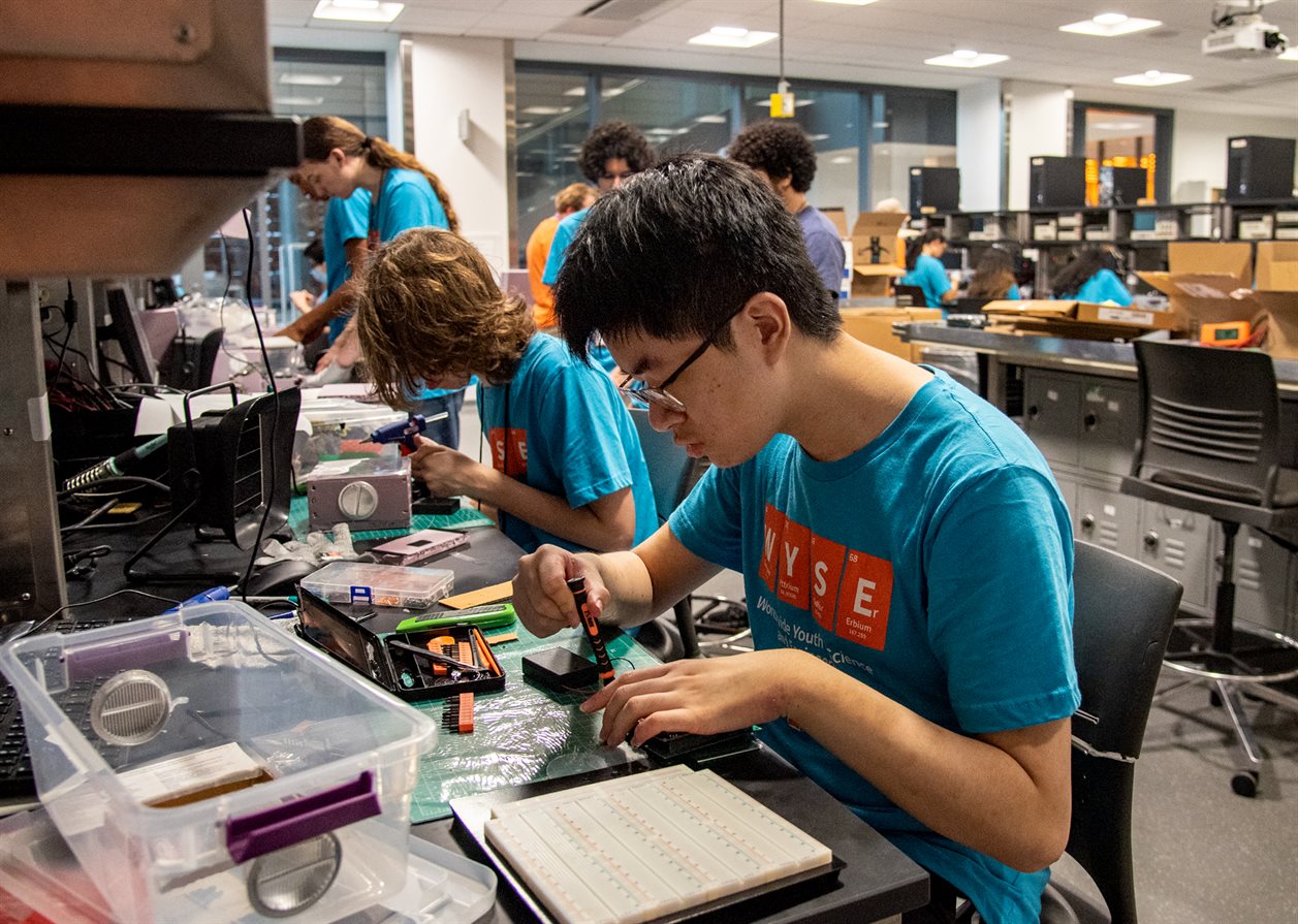 High school students in turquoise t-shirts are leaning over a counter in a lab, working.