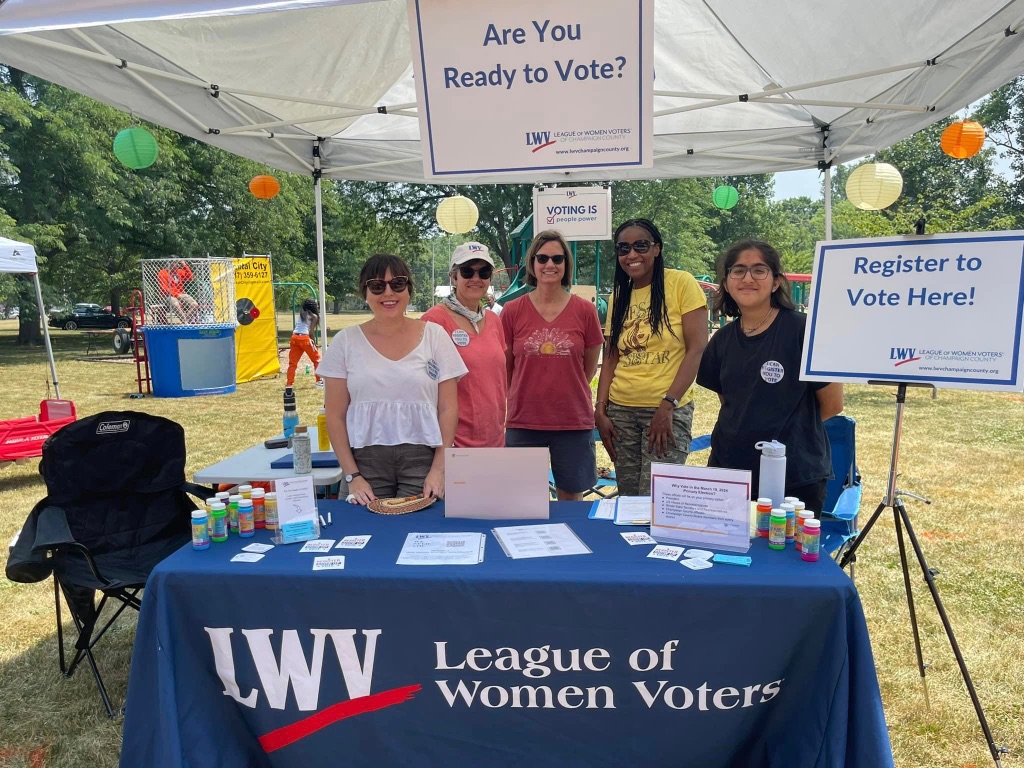 A group of women stand together at a table with a blue League of Women voters printed on it in white letters. They are standing behind the information table that has fliers and brochures. The woman on the left is white with brown hair, the next woman is white with a pink shirt and white hat, the third is a white woman in a maroon shirt, the fourth woman is black with medium length dark brown braids and a yellow t-shirt, the woman on the right end is brown with shorter brown hair and a black t-shirt with a pin that says "you can vote".