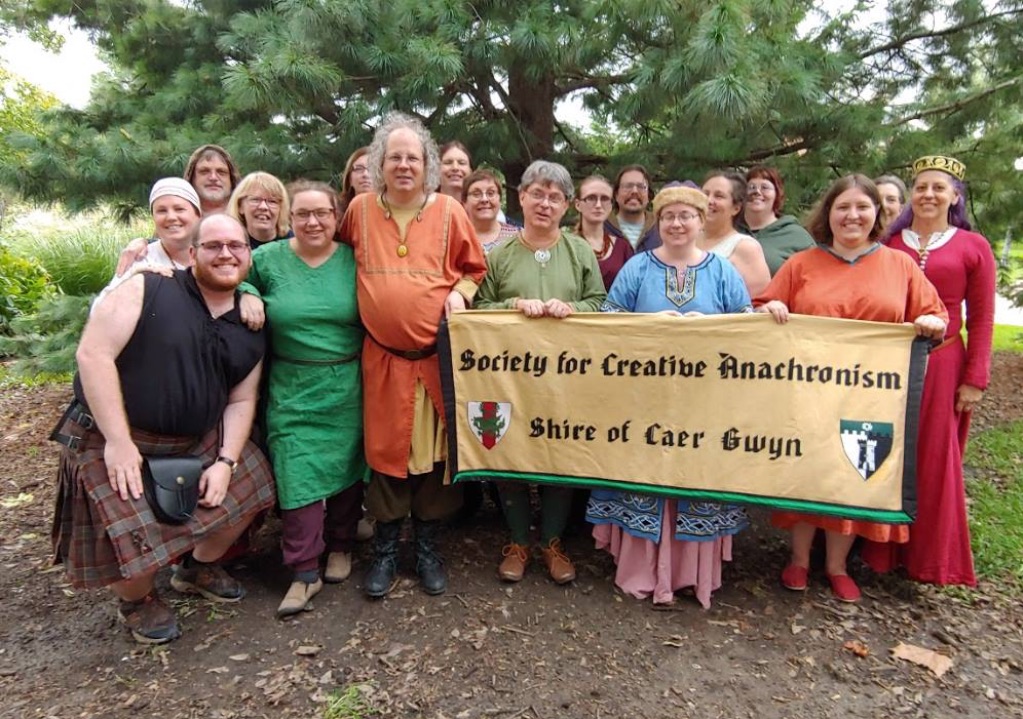 a group of white people dressed in medieval clothing stand together looking at the camera. They are holding a large sign that says Society for Creative Anachronism Shire of Care Gwyn.