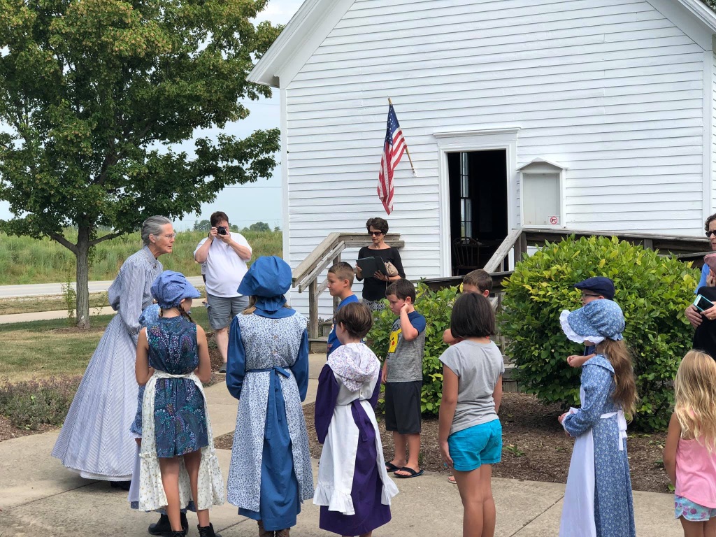 a white woman dressed in a long light blue dress and bonnet acts as a school teacher from the early 18th century. She is standing in front of a simple white building and a group of children, some also dressed in costumes others wearing shorts and a t-shirt.