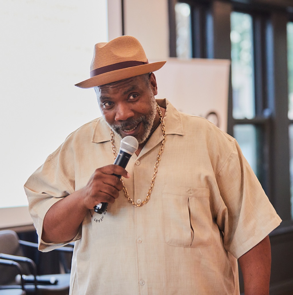A black man with a graying beard, wears a tan collared shirt with a front pocket and a straw hat with a brim. He is holding a microphone up to his mouth and looking off to the right.