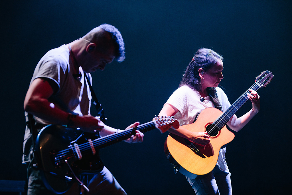 The image showcases two musicians on a stage, bathed in blue light. The musician on the left is playing an electric bass guitar, while the one on the right is strumming an acoustic guitar. The background is dark, emphasizing the musicians and their instruments.