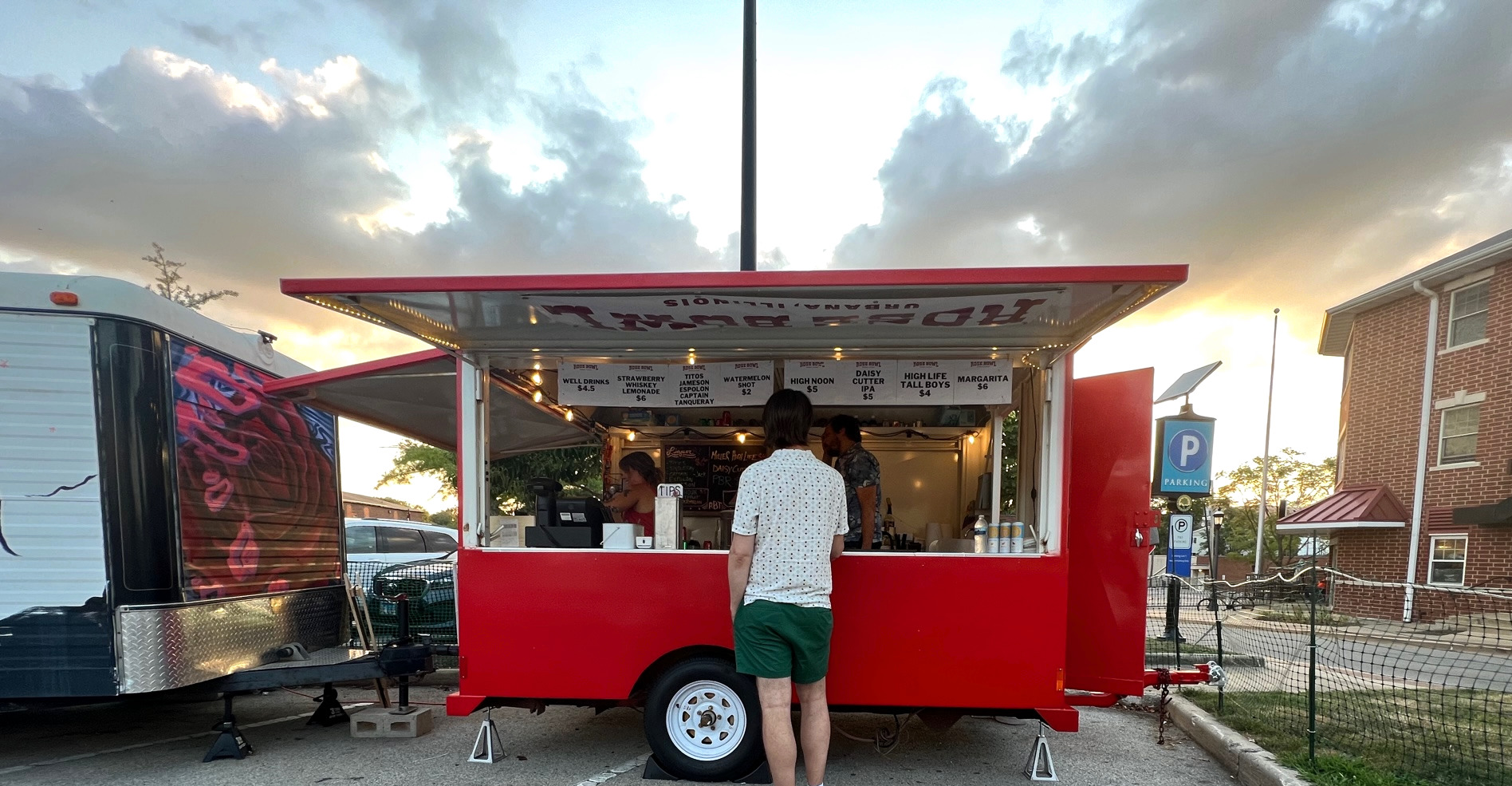 A red food truck in the parking lot of the Rose Bowl Tavern's outdoor space. A man looks at the menu of drinks.
