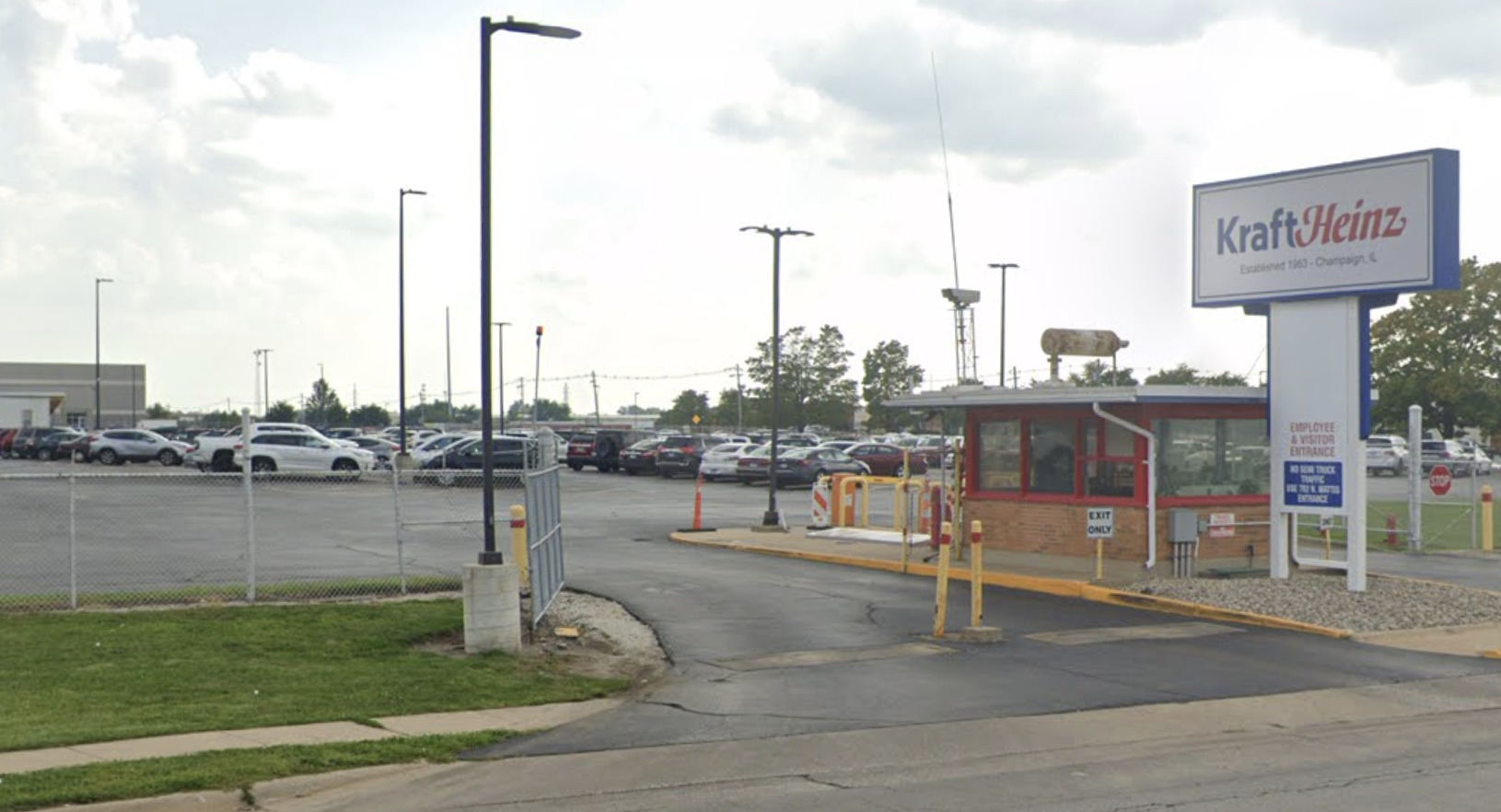 The entrance for the Kraft Heinz factory in Champaign, IL. A security station with brick foundation and red trim sits behind a large white and blue sign the reads KraftHeinz. Beyond the entrance is a large parking lot with many cars parked.