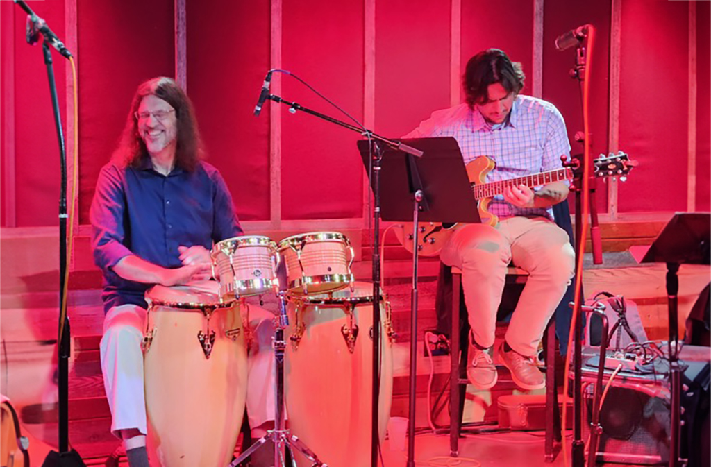 A stage with a red background hosts two musicians. One is engaged with a set of conga drums, while the other strums an electric guitar. Both are seated on stools, with music stands before them. A microphone stand graces the right side of the stage. The floor beneath them is wooden.