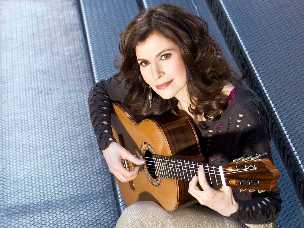 I brunette woman playing guitar while kneeling on steel flooring looking up at the camera.