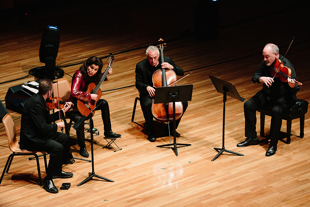 The image presents a string quartet performing on a stage. The quartet is composed of two violinists, a cellist, and a violist. They are seated on wooden chairs, each playing their respective instrument. Music stands in front of them hold their sheet music. The stage features a wooden floor and a black curtain as the backdrop. Two speakers flank the stage on either side.
