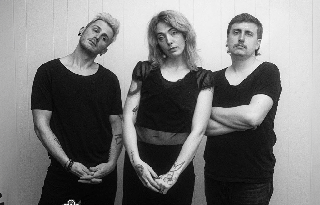 Three individuals are standing in front of a white wall. All are dressed in black t-shirts. The individual in the center appears to be a woman with tattoos on her arms and stomach. The individual on the left has his arms crossed, while the one on the right has his arms folded. The photo is in black and white.
