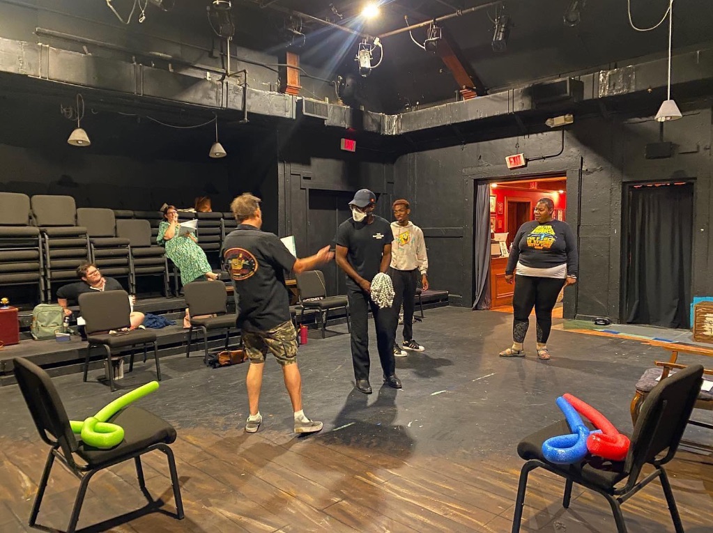 A group of people stand on a stage  rehearsing a play. The walls and floor are black. There are black chairs in the foreground with long balloon swords in red, blue, and green. 